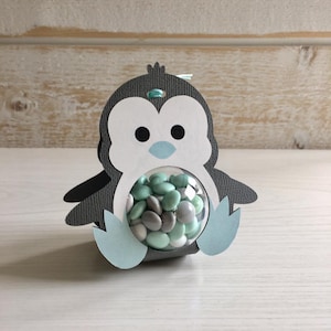 May include: A gray and white penguin-shaped paper party favor with a clear plastic sphere filled with small green and gray candies.