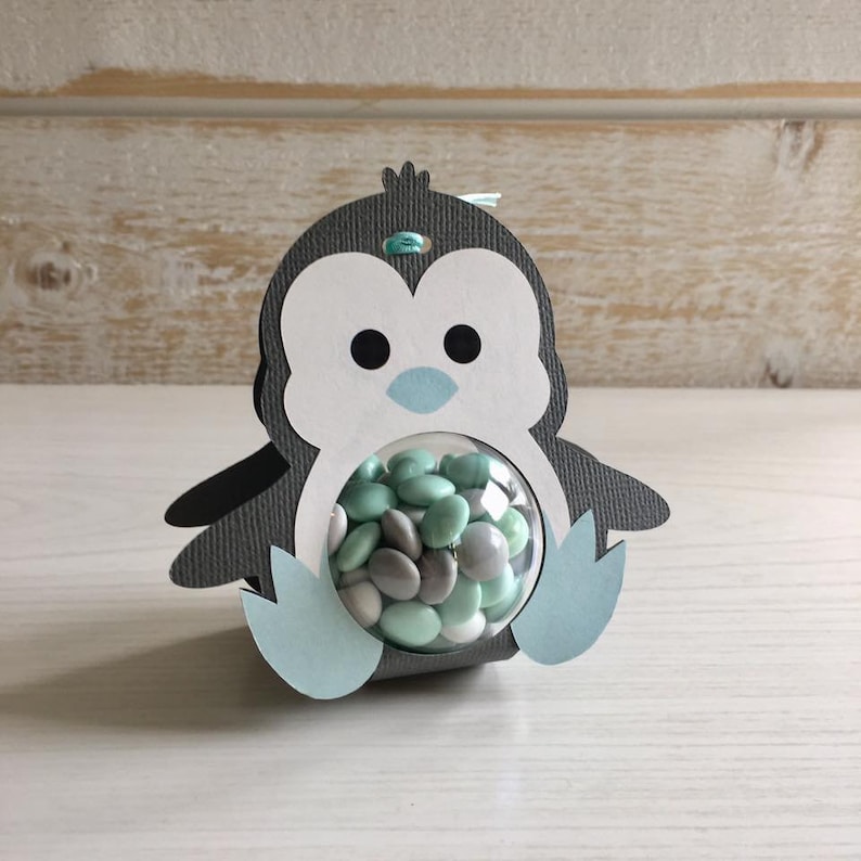 May include: A gray and white penguin-shaped paper party favor with a clear plastic sphere filled with small green and gray candies.