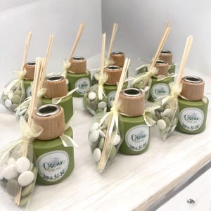 Op de afbeelding: Groene glazen flessen met houten deksels en een zilveren ring, gevuld met witte en groene snoepjes. Elke fles heeft een etiket met de naam "Oscar" en een datum. De flessen zijn gebonden met een doorschijnend geel lint en hebben houten stokjes die uit de bovenkant steken.