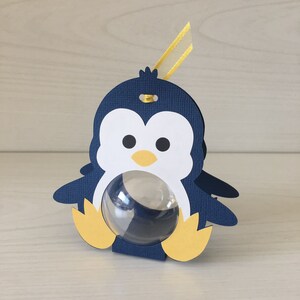 May include: A blue penguin-shaped favor box with a clear plastic window. The box has yellow feet and a yellow ribbon.