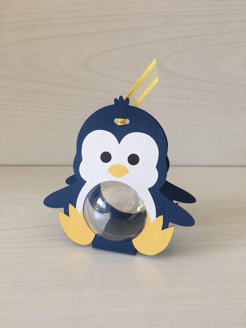 May include: A blue penguin-shaped favor box with a clear plastic window. The box has yellow feet and a yellow ribbon.