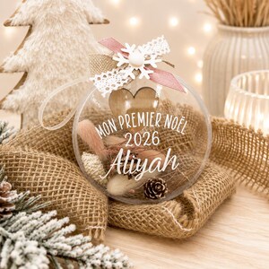 Puede incluir: Un adorno navideño esférico y transparente relleno de flores secas, una pequeña piña y un corazón de madera. El frente presenta el texto blanco MON PREMIER NOEL 2026 Aliyah. Está rematado con un copo de nieve blanco, encaje y cinta rosa.
