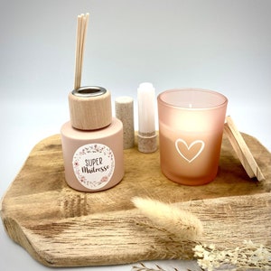 Op de afbeelding: Een cadeauset met een roze diffuserfles met een houten dop en een label met de tekst "SUPER Maitresse", een roze kaars met een wit hartontwerp, een kleine witte tube en een houten deksel.