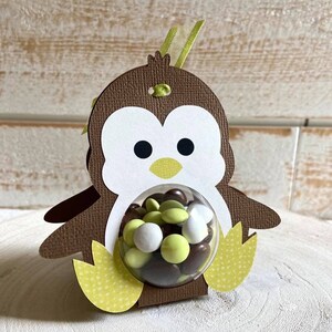 May include: A brown penguin-shaped cardboard favor box with a clear plastic window. The box is filled with chocolate and green candy. The penguin has a yellow beak and feet.