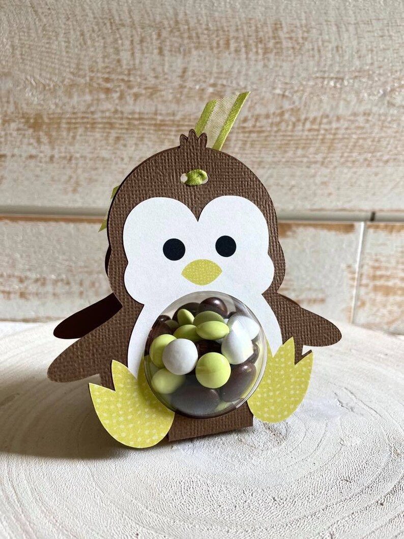 May include: A brown penguin-shaped cardboard favor box with a clear plastic window. The box is filled with chocolate and green candy. The penguin has a yellow beak and feet.