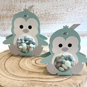 May include: Two light blue penguin-shaped favor boxes with clear plastic domes filled with pastel green and gray candy. The boxes are made of cardstock and have a gray ribbon tied around the top.