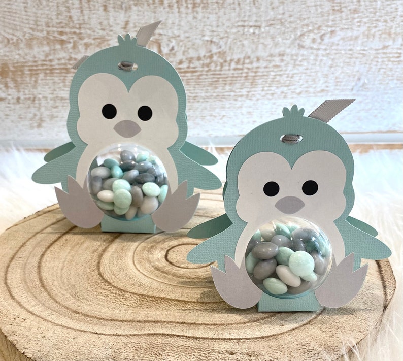 May include: Two light blue penguin-shaped favor boxes with clear plastic domes filled with pastel green and gray candy. The boxes are made of cardstock and have a gray ribbon tied around the top.