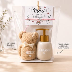 Op de afbeelding: Een cadeau set in een doorzichtige zak, inclusief een kleine teddybeer met een strik, een lotiondispenser en een kaart met de tekst "Merci". De teddybeer heeft het woord "Merci" erop. De lotiondispenser heeft het woord "Madame" erop.