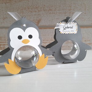 May include: Two gray penguin-shaped favor boxes with clear plastic windows. The boxes have yellow feet and beaks. One box has a tag that says "Gabriel 21 September 2019".
