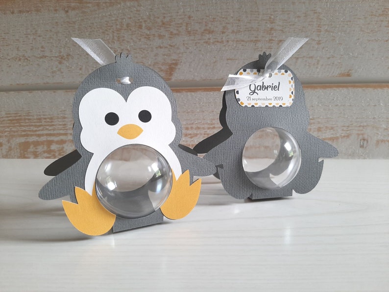 May include: Two gray penguin-shaped favor boxes with clear plastic windows. The boxes have yellow feet and beaks. One box has a tag that says "Gabriel 21 September 2019".