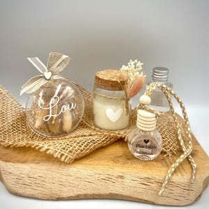 Op de afbeelding: Een set van drie decoratieve glazen ornamenten met gedroogde bloemen en een hartbedel. De ornamenten liggen op een houten oppervlak met jute stof. Een ornament zegt "Lou" en een ander zegt "Marraine d'Amour".