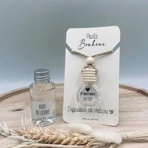 Op de afbeelding: Een witte kaart met de tekst "Porte Bonheur" en een klein glazen flesje met een ketting van houten kralen. Het flesje is gelabeld met "Parrain Au Top" en "Diffuseur de voiture". Het flesje is gevuld met een heldere vloeistof en heeft een zilveren dop. De kaart ligt op een houten oppervlak met een tarwehalm.