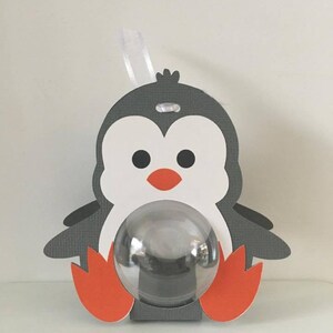May include: A gray and white penguin-shaped favor box with a clear plastic window. The penguin has orange feet and a red beak. The box has a white ribbon attached to the top.