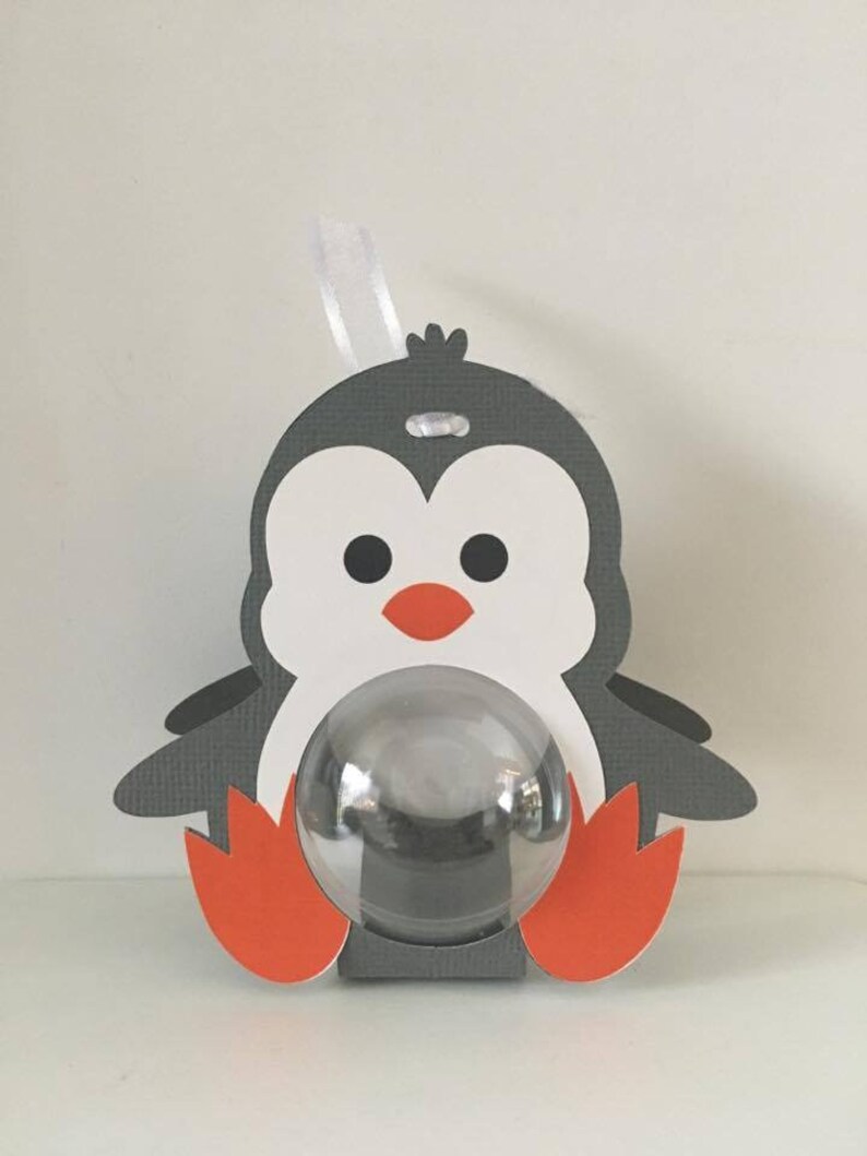 May include: A gray and white penguin-shaped favor box with a clear plastic window. The penguin has orange feet and a red beak. The box has a white ribbon attached to the top.