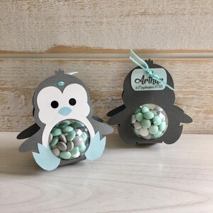 May include: Two gray penguin-shaped party favor boxes with clear plastic windows. The boxes are filled with small, round candies in mint green and white. One box has a tag attached that says "Arthur le 1 septembre 2018".