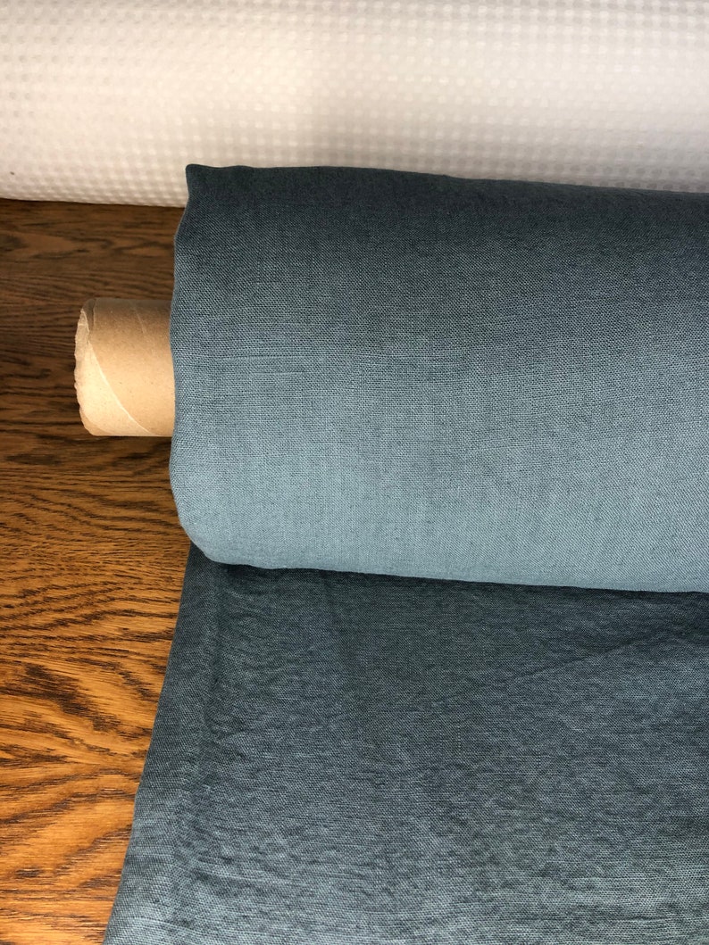 May include: A roll of blue linen fabric on a wooden surface. The fabric has a subtle texture and a slightly darker shade at the edges.