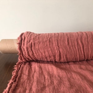 Puede incluir: Un rollo de tela de lino de color rosa polvoriento con bordes sin rematar. La tela está enrollada en un tubo de cartón.