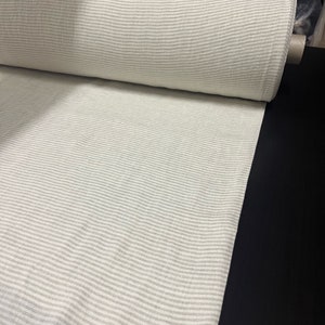 Może przedstawiać: Rolka białej tkaniny w cienkie szare paski. Tkanina jest złożona na pół, a rolka leży na czarnej powierzchni.