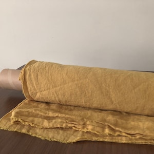 Könnte beinhalten: Eine Rolle gelber Leinenstoff mit einem rohen Saum. Der Stoff ist aufgerollt und mit einem braunen Papierband gebunden.