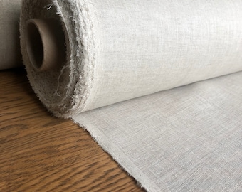 May include: A roll of natural linen fabric, partially unrolled, revealing a textured weave. The fabric is a light, neutral color, with a raw, unfinished edge. The roll sits on a wooden surface, showcasing the fabric's drape and texture.