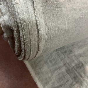 Può includere: Un rotolo di tessuto grigio chiaro con una superficie leggermente strutturata. Il tessuto ha un bordo grezzo ed è arrotolato strettamente.