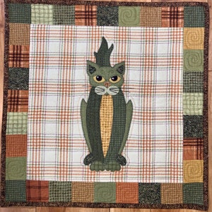 May include: A black and green cat applique on a white and brown checked fabric background. The cat is standing on its four paws with its tail up. The fabric is surrounded by a brown border with a checked pattern.