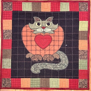 May include: A quilted wall hanging featuring a stylized cat with a heart in its chest. The cat is surrounded by a plaid pattern in shades of red, orange, and brown. The background is a dark brown fabric with a grid pattern.