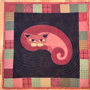 Op de afbeelding: Een rood gestikt kattenontwerp op een zwarte achtergrond met een rode, groene en bruine geruite rand.