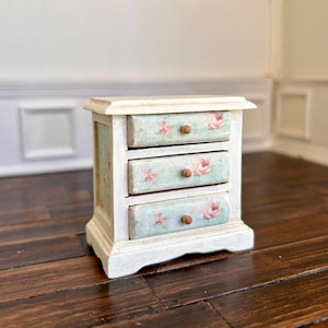 Może przedstawiać: Mała, drewniana komoda z trzema szufladami i postarzanym białym wykończeniem. Szuflady zdobione są kwiatowym wzorem w kolorze różowym i zielonym, z małymi drewnianymi gałkami. Dekoracyjny element.