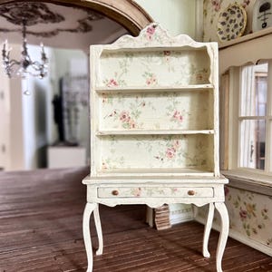 Op de afbeelding: Een witte miniatuurkast met drie planken en een lade. De binnenkant en achterkant van de kast zijn versierd met een bloemmotief van roze rozen. De kast heeft gebogen poten en houten knoppen.