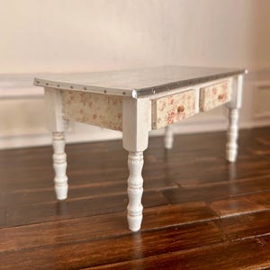 Pode incluir: Uma mesa branca em miniatura com duas gavetas e pernas torneadas. A mesa tem um tampo de metal e laterais e frentes de gavetas com padrão floral. As gavetas têm pequenos puxadores de madeira. A mesa tem cerca de 30 cm de comprimento.