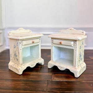 Może przedstawiać: Dwa miniaturowe kremowe stoliki nocne z postarzanym wykończeniem. Każdy ma małą szufladę z drewnianym uchwytem i otwartą półkę. Niebieskie kwiatowe akcenty zdobią boki. Stoliki nocne są umieszczone na ciemnej drewnianej powierzchni.