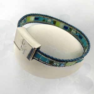Peut inclure: Un bracelet en cuir bleu avec un fermoir argenté et un motif en mosaïque multicolore. La mosaïque présente des carreaux turquoise, bleus, verts et blancs.