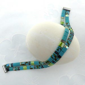 Peut inclure: Un collier de perles turquoise avec un fermoir magnétique. Le collier présente des perles carrées avec des accents de vert, de noir et d'argent. Le collier est drapé sur une pierre blanche de forme ovale.