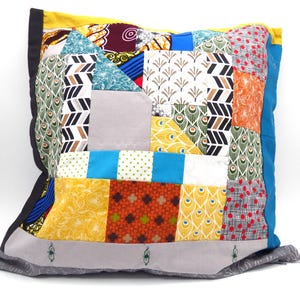 May include: Square patchwork pillow with a variety of colorful fabric patterns. The pillow features a mix of geometric shapes, floral prints, and peacock feather designs in shades of blue, yellow, orange, and red. The edges are trimmed with black and gray fabric.