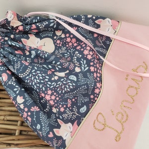 Op de afbeelding: Een roze en marineblauwe trekkoordtas met een bloemen- en reeënpatroon. De tas heeft een roze paneel met gouden glitterletters en roze linten. De tas staat in een rieten mand.