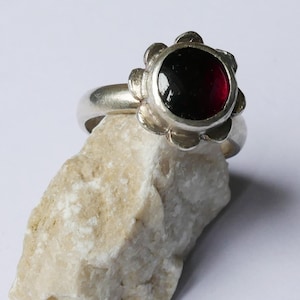 Può includere: Un anello d'argento con una montatura a forma di fiore che tiene una gemma rotonda di colore rosso scuro. L'anello è esposto su una pietra chiara e strutturata su sfondo bianco. La gemma è di un rosso intenso.