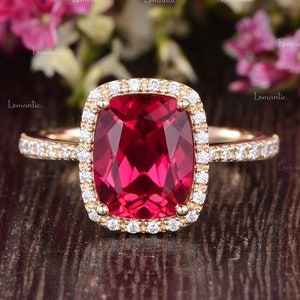 May include: A gold ring featuring a large, cushion-cut ruby encircled by a halo of smaller diamonds. The ring is displayed on a wooden surface with pink flowers in the background.