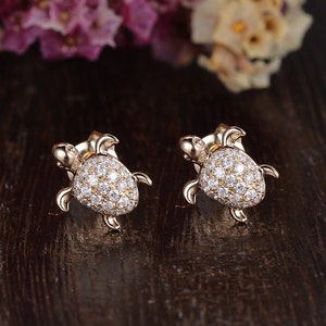 May include: A pair of gold-toned earrings shaped like turtles, each turtle is encrusted with small, clear cubic zirconia stones.