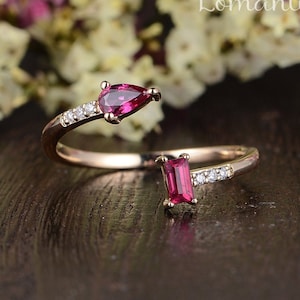 May include: A rose gold ring with two ruby stones. The ring is designed to be worn on the middle finger. The ruby stones are a pear shape and a rectangular shape. The ring is set with small diamonds.