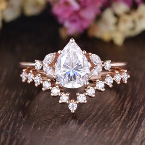 Könnte beinhalten: Ein Verlobungsring mit einem birnenförmigen Diamanten in Roségold mit einem zarten Band aus kleineren Diamanten. Der Ring liegt auf einer Holzoberfläche.