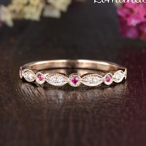 Puede incluir: Una delicada banda de oro rosa con una fila de pequeños diamantes y piedras preciosas rojas. La banda está colocada sobre una superficie de madera oscura.