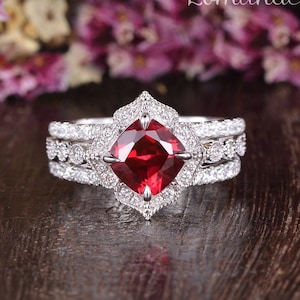 Peut inclure: Une bague de fiançailles en trois parties avec une grande pierre rouge de forme carrée entourée de petites pierres claires. La bague est sertie d'argent.