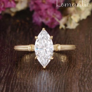 May include: A gold ring featuring a large, clear, marquise-cut center stone held by six prongs. The band has a distinct ribbed, multi-line texture that extends horizontally from the setting. The word Lomantic is visible in the upper right corner.