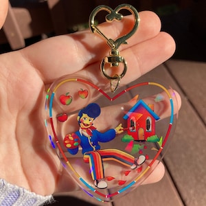 May include: Heart-shaped keychain with a colorful illustration of a figure painting a red house. The keychain has a gold-colored clasp. The illustration includes apples and rainbow-striped pants.