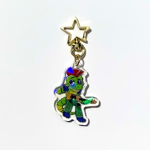 May include: A green and red cartoon alligator with a mohawk playing a guitar. The alligator is wearing green and black plaid trousers. The image is on a clear acrylic keychain with a gold star charm.