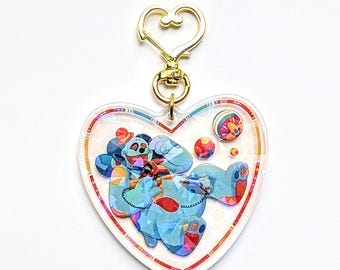 Può includere: Un portachiavi in ​​acrilico trasparente a forma di cuore con un'illustrazione colorata di un personaggio blu con un cappello rosso e una camicia a righe rosse e bianche. Il personaggio è circondato da palle colorate e una catena dorata.