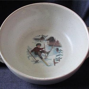 May include: A white ceramic serving bowl with a speckled glaze and a winter scene illustration of a person skiing. The scene includes a small cabin and trees.