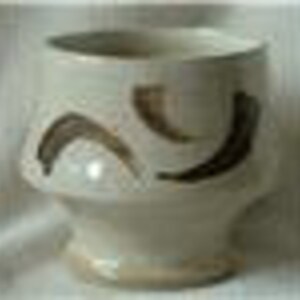 May include: A white ceramic cup with a brown glaze and a unique design of curved lines. The cup has a wide base and a rounded top.