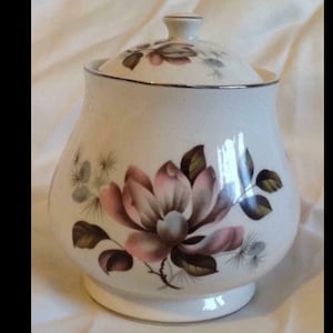 May include: White ceramic jar with a lid, featuring a floral design with pink and brown flowers. The jar has a gold trim around the lid and base.